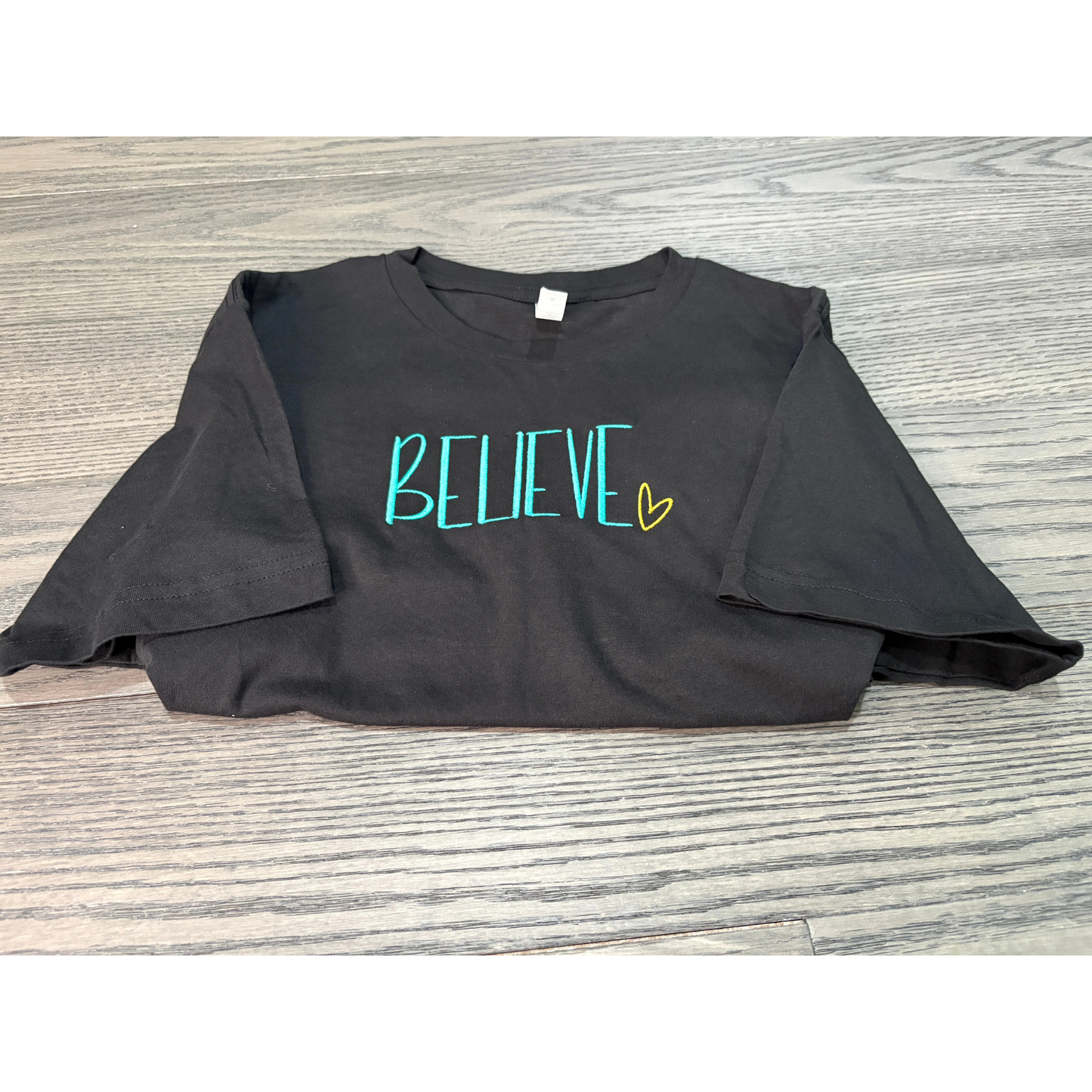 Black t-shirt with 'BELIEVE' printed on it in teal and yellow, laid flat on a wooden surface.