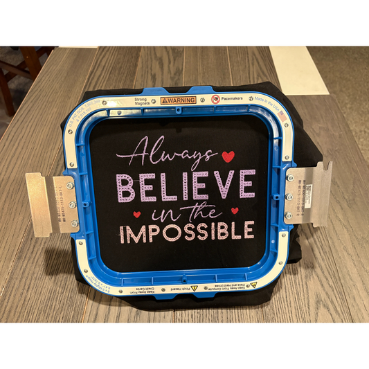 Black shirt with 'Always Believe in the Impossible' text on a wooden surface inside an embroidery hoop
