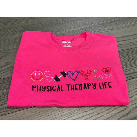 Folded pink t-shirt with 'Physical Therapy Life' text and symbols on a wooden surface