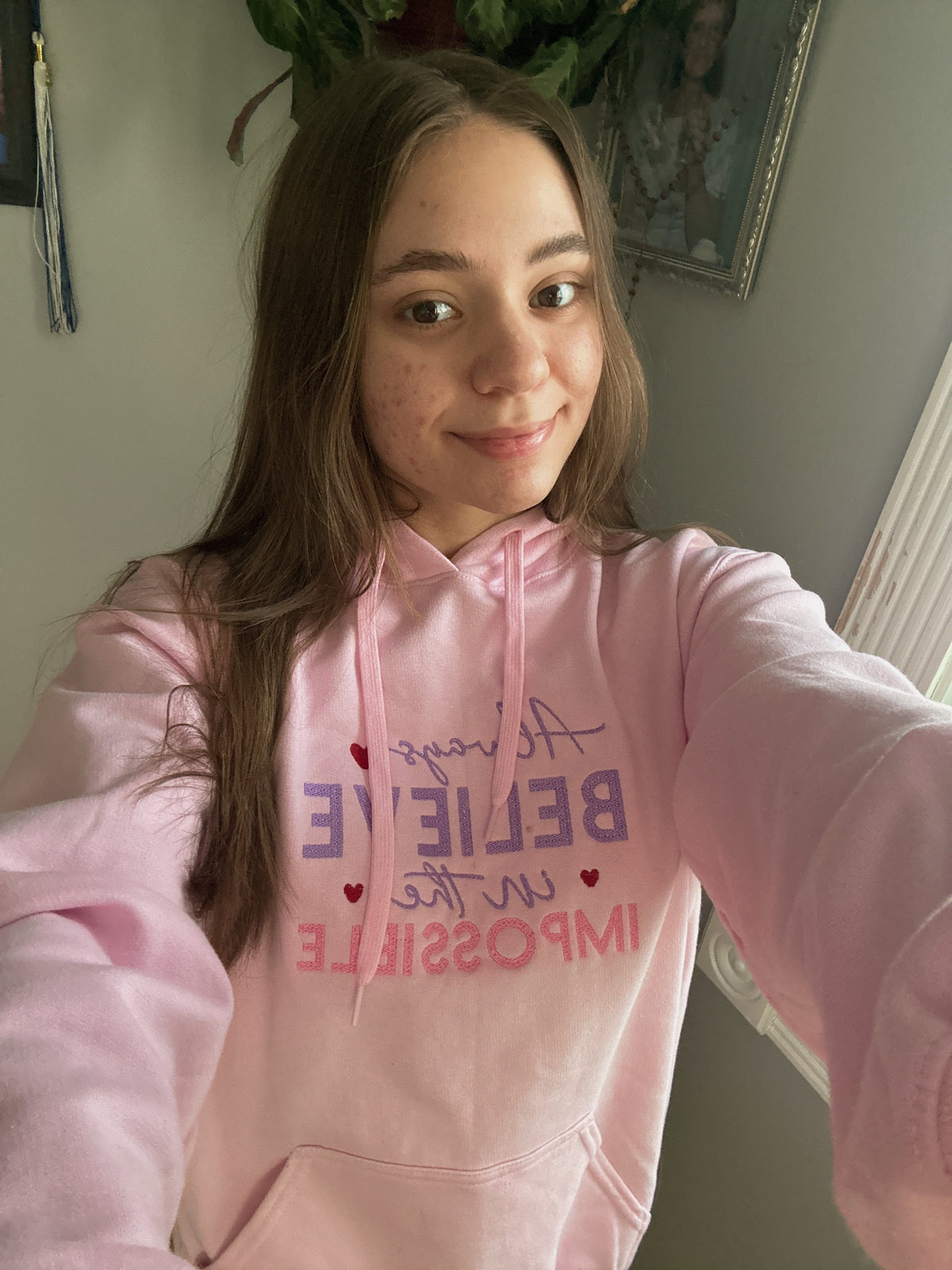 Person wearing a pink hoodie with motivational text, Always believe in the Impossible, taking a selfie.