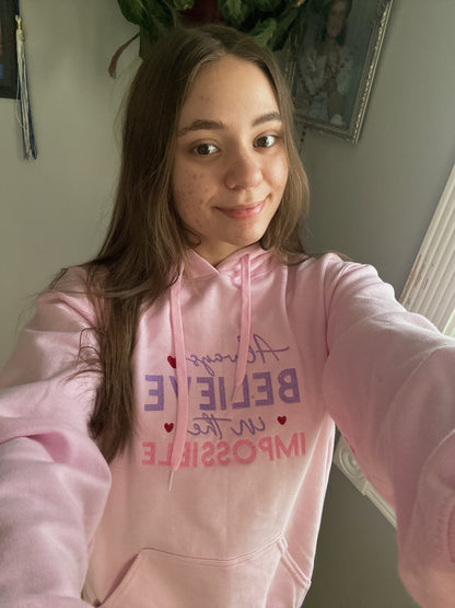 Person wearing a pink hoodie with motivational text, Always believe in the Impossible, taking a selfie.