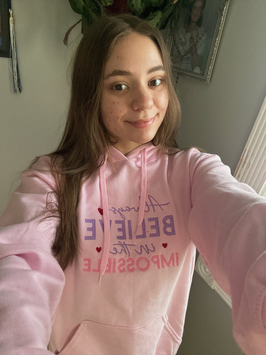 Person wearing a pink hoodie with motivational text, Always believe in the Impossible, taking a selfie.