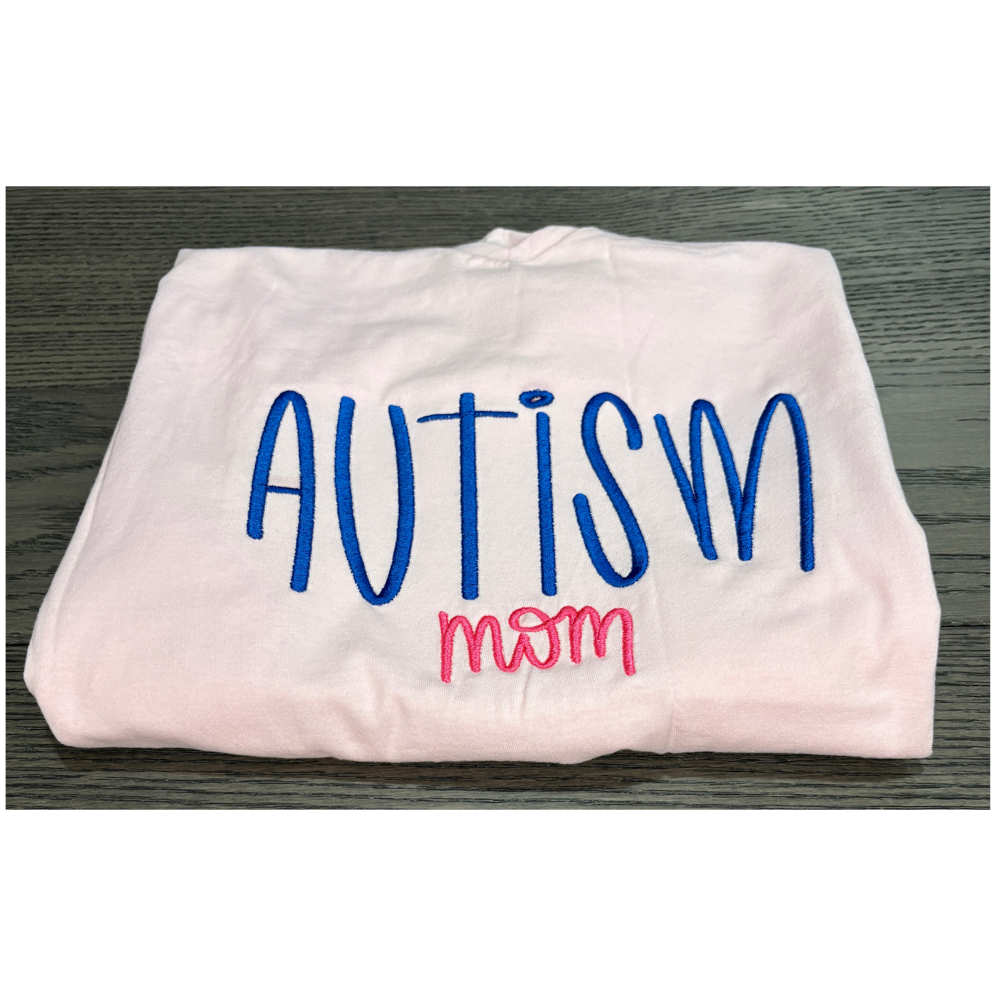 Folded white shirt with 'Autism mom' text in blue and pink on a wooden surface