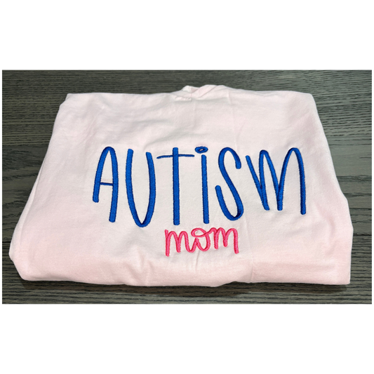 Folded white shirt with 'Autism mom' text in blue and pink on a wooden surface