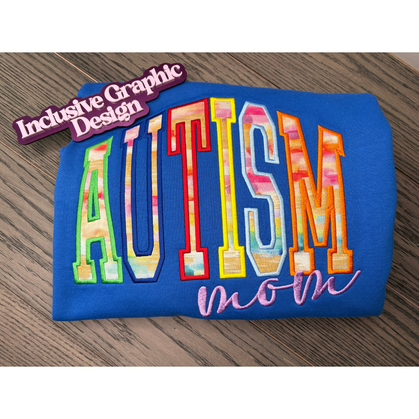 A folded blue sweatshirt on a wooden surface, featuring embroidered text that reads 'AUTISM mom' in multi  brushed colored fabric with a purple text reading 'mom' underneath. A purple magnet on the top left reads 'Inclusive Graphic Design'.