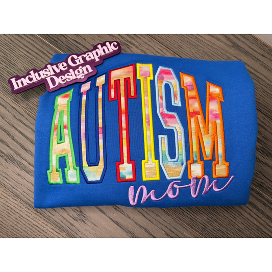 A folded blue sweatshirt on a wooden surface, featuring embroidered text that reads 'AUTISM mom' in multi  brushed colored fabric with a purple text reading 'mom' underneath. A purple magnet on the top left reads 'Inclusive Graphic Design'.