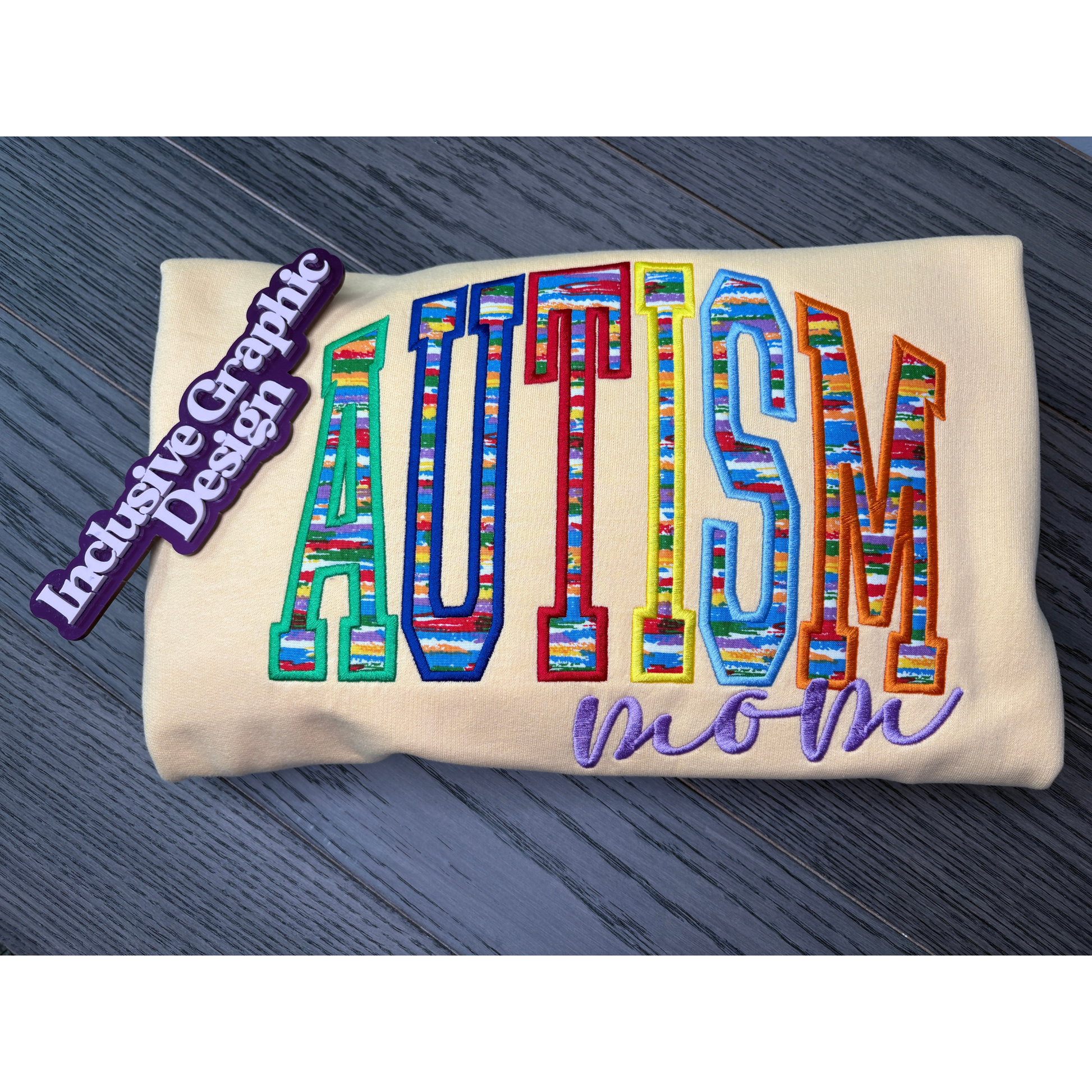 A folded cream-colored sweatshirt, featuring colorful embroidered text that reads 'AUTISM' with a patterned fill fabric and 'mom' in purple script below. A purple magnet on the left reads 'Inclusive Graphic Design'.