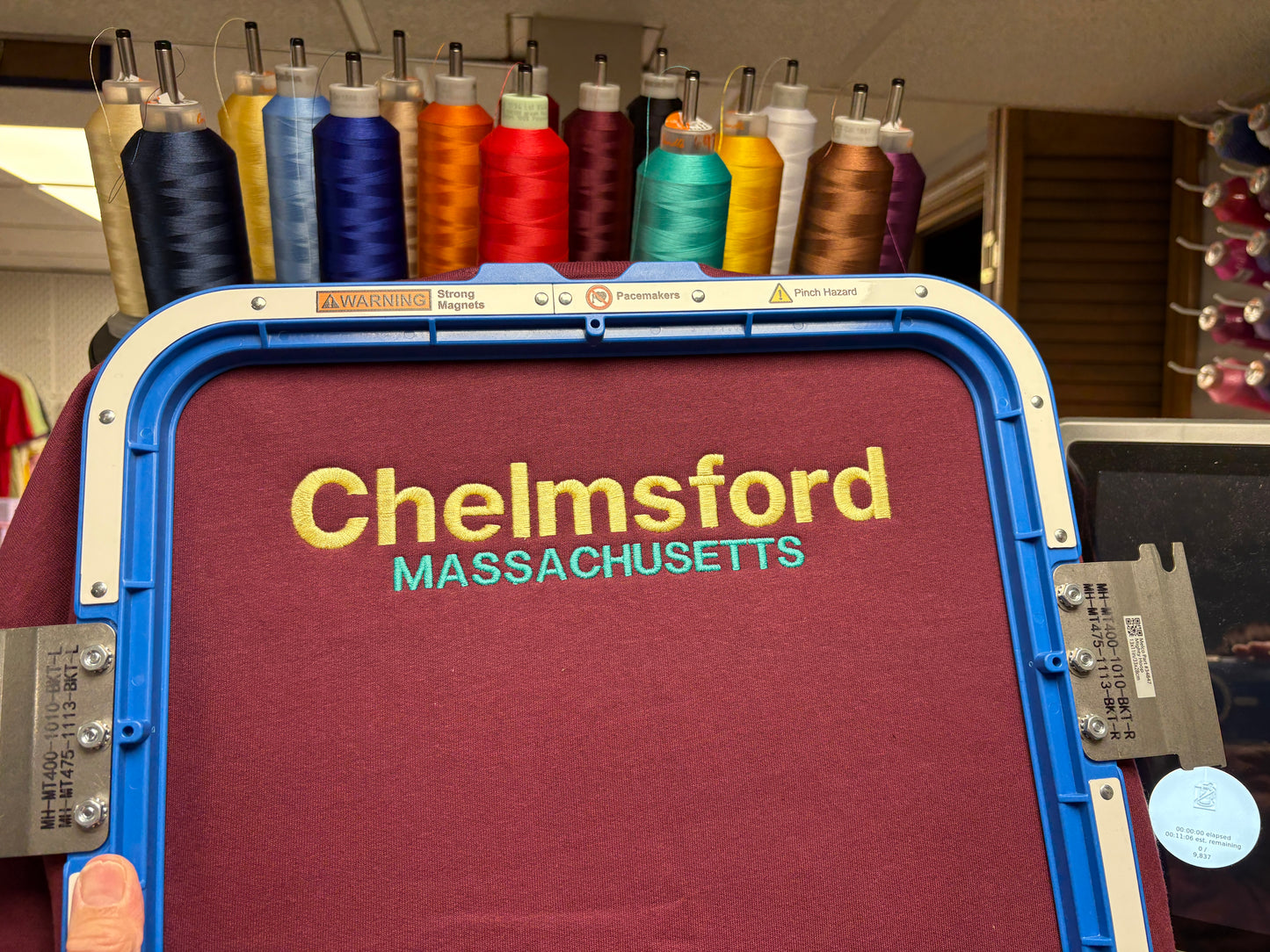 A close-up of an embroidery machine hoop holding a burgundy tshirt with the words "Chelmsford" in yellow and "MASSACHUSETTS" in teal embroidered on the fabric. Spools of thread in many colors are visible in the background.