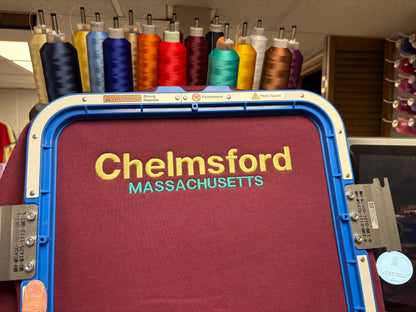 A close-up of an embroidery machine hoop holding a burgundy tshirt with the words "Chelmsford" in yellow and "MASSACHUSETTS" in teal embroidered on the fabric. Spools of thread in many colors are visible in the background.