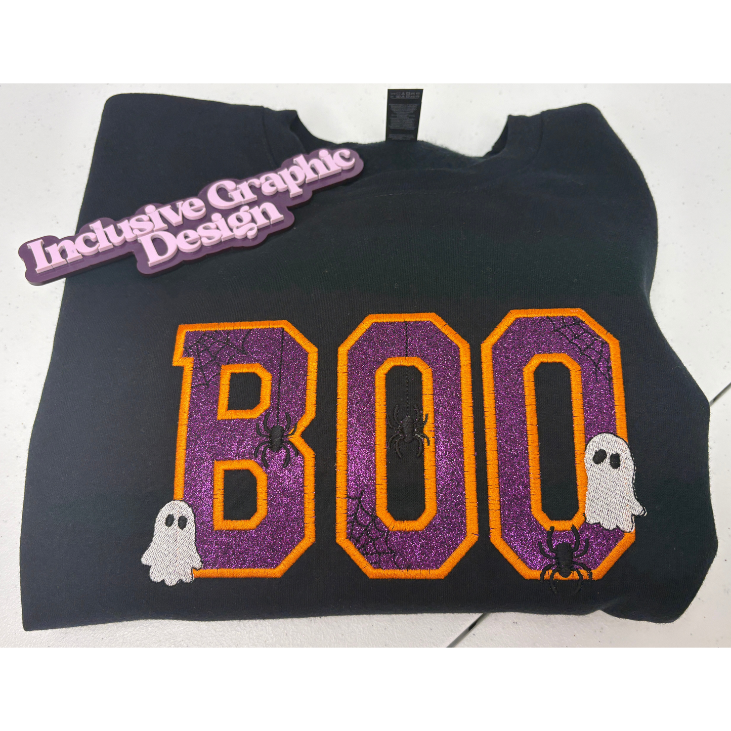 A black sweatshirt featuring a large 'BOO' graphic in purple glitter with orange borders, decorated with small spider and ghost motifs. A purple 'Inclusive Graphic Design' brand tag is attached near the collar.