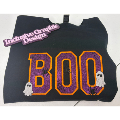 A black sweatshirt featuring a large 'BOO' graphic in purple glitter with orange borders, decorated with small spider and ghost motifs. A purple 'Inclusive Graphic Design' brand tag is attached near the collar.