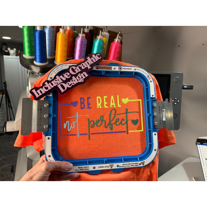 Orange t-shirt with 'Be Real, Not Perfect' text in a graphic design machine.