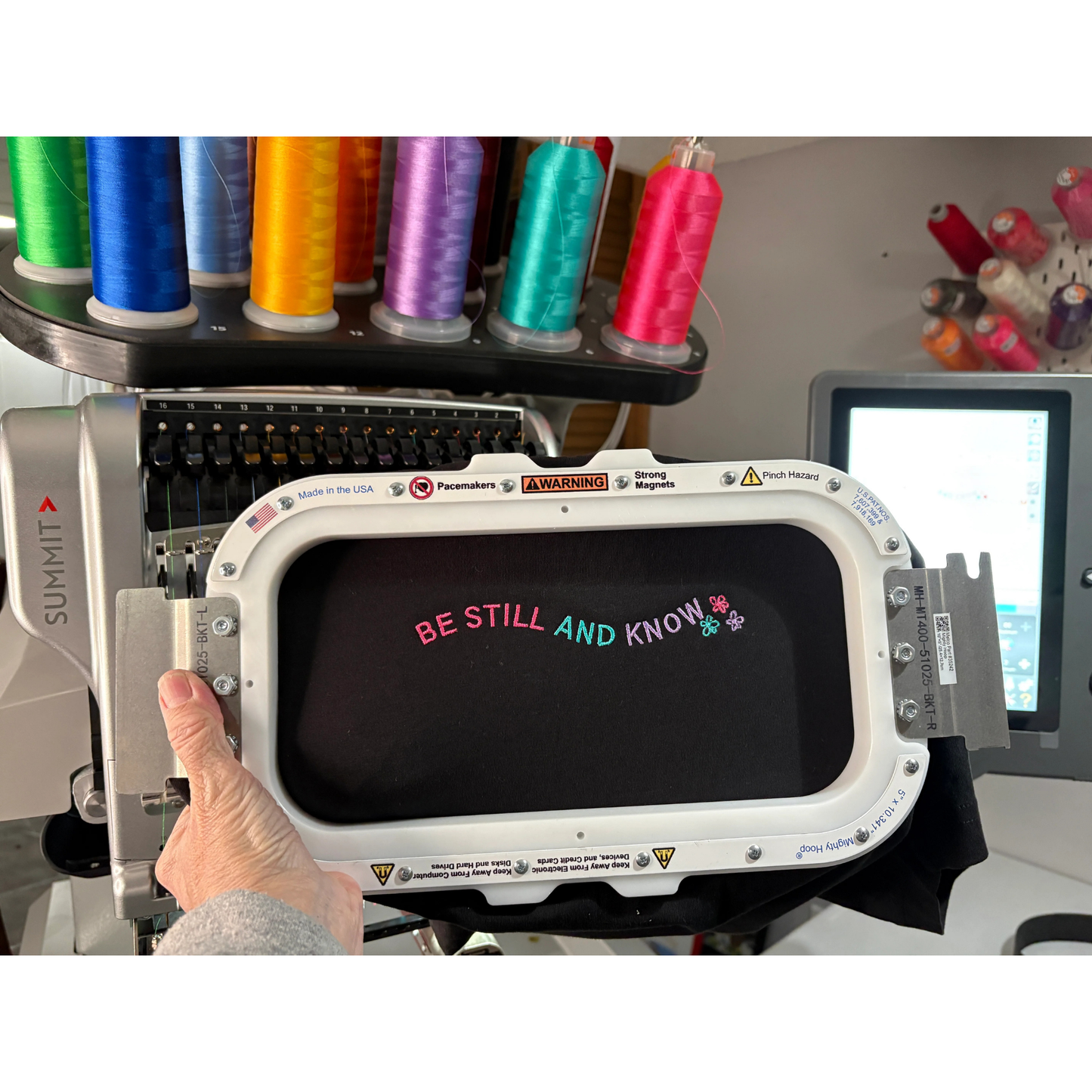 Embroidery machine with colorful thread spools and a digital display showing 'Be Still and Know'.