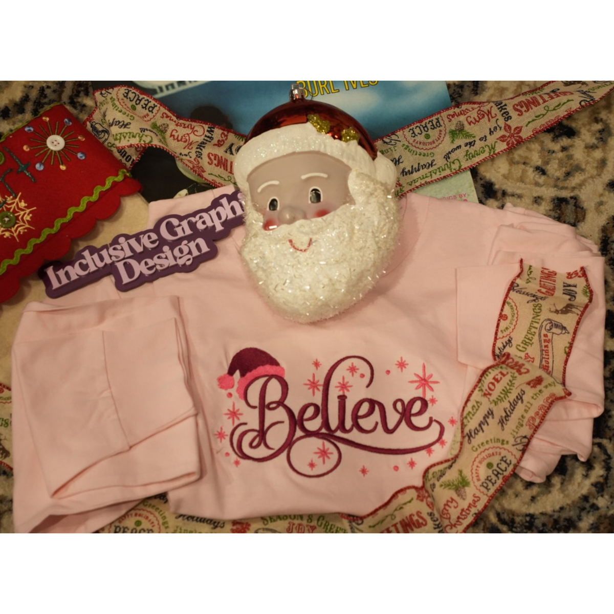 A folded light pink long-sleeve shirt with a glittery white Santa beard decoration and a red 'Believe' graphic, displayed on a patterned fabric background with a Santa hat and ribbon.