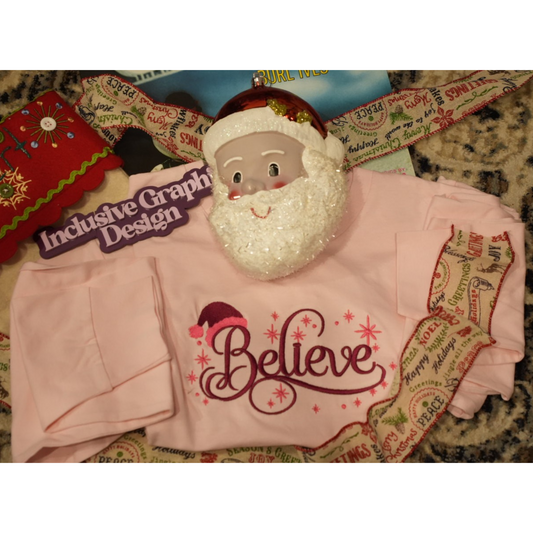 A folded light pink long-sleeve shirt with a glittery white Santa beard decoration and a red 'Believe' graphic, displayed on a patterned fabric background with a Santa hat and ribbon.