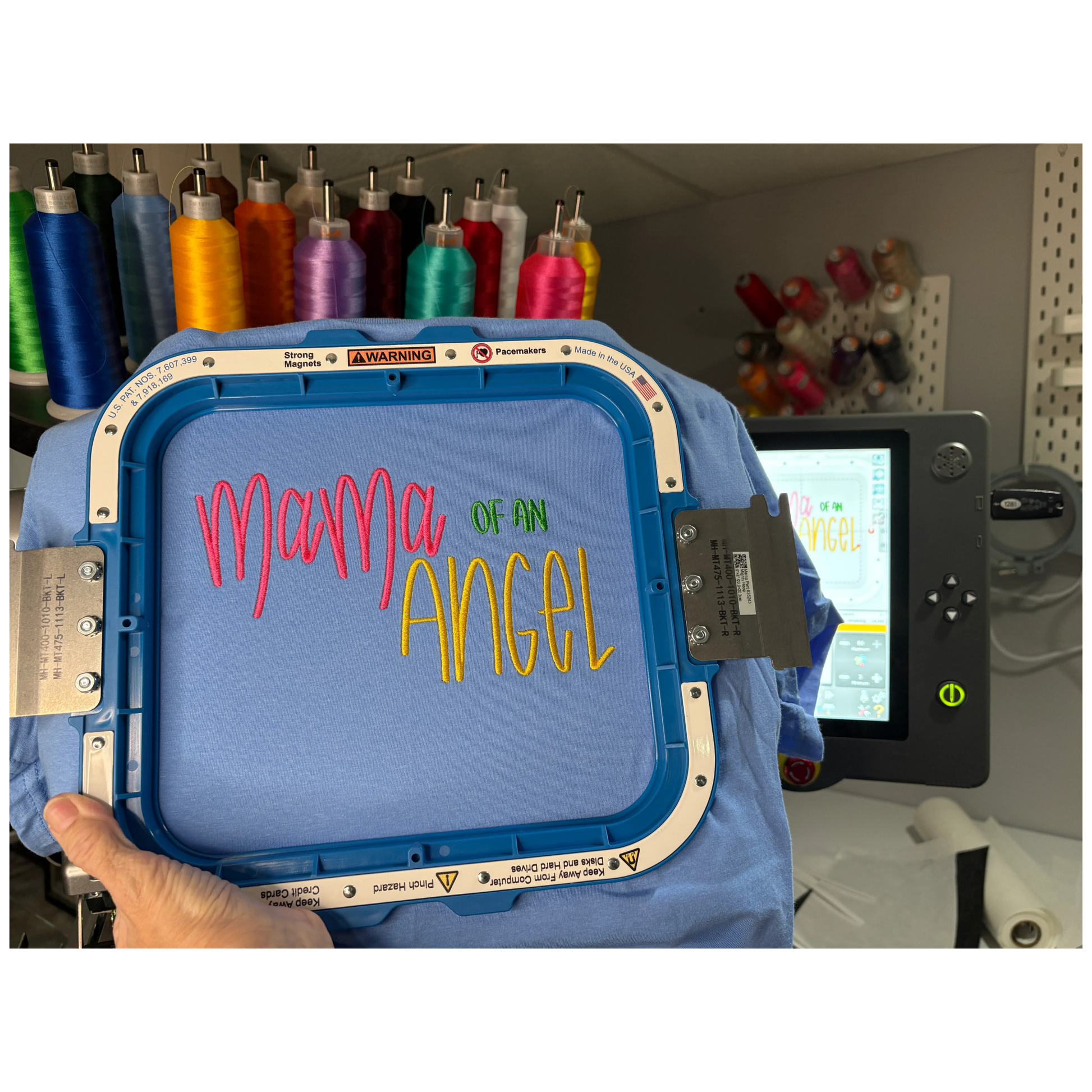 Embroidery machine hoop with 'Mama of an Angel' text in colorful letters, surrounded by spools of thread.