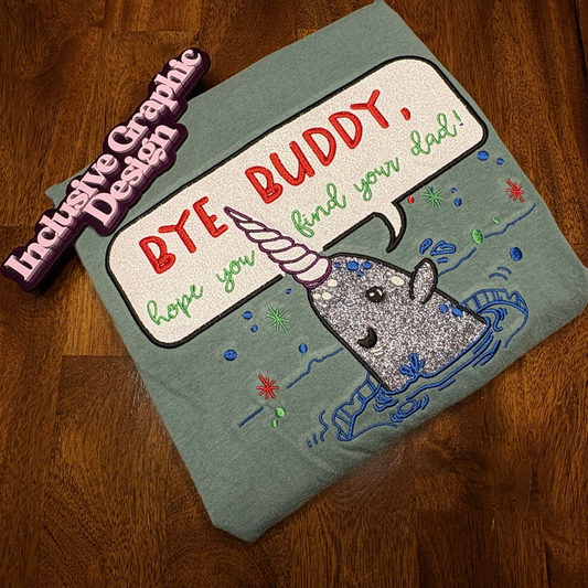 A folded sage green t-shirt with a glittery gray narwhal graphic and a speech bubble reading 'BYE BUDDY, hope you find your dad!' in red and green text. A 3D purple and pink 'Inclusive Graphic Design' sign is propped up next to it on a dark wood floor.