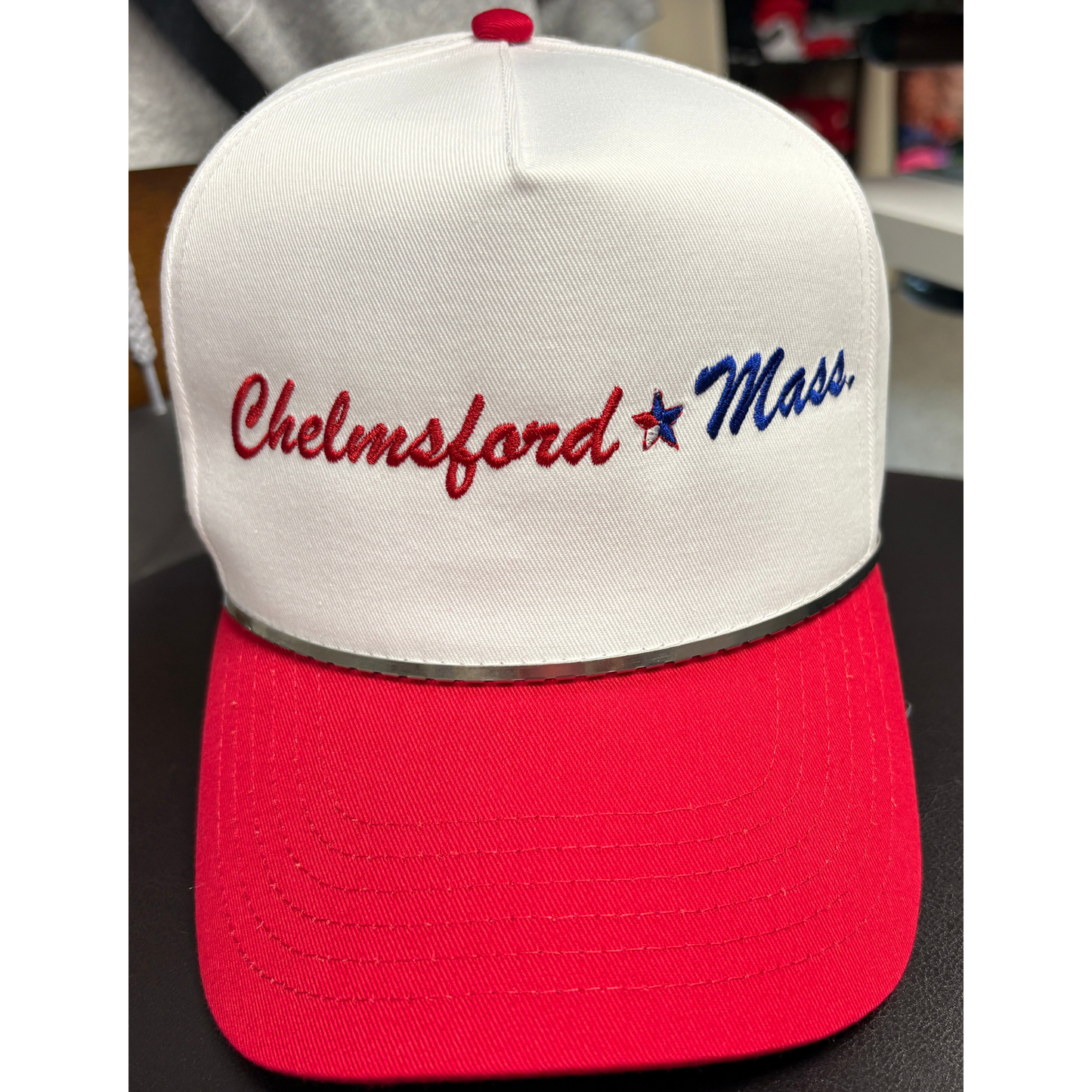 a white hat with a red brim with embroidered text chelmsford in red and Mass in blue with a red, white and blue star seperating the 2 texts. 