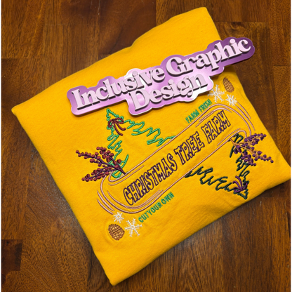 A folded bright yellow t-shirt with colorful embroidered graphics on a dark wood background. The embroidery features a Christmas tree, pinecones, and the text 'CHRISTMAS TREE FARM' and 'cut Your Own, Farm Fresh'. A purple magnet with the words Inclusive Graphic Design on top