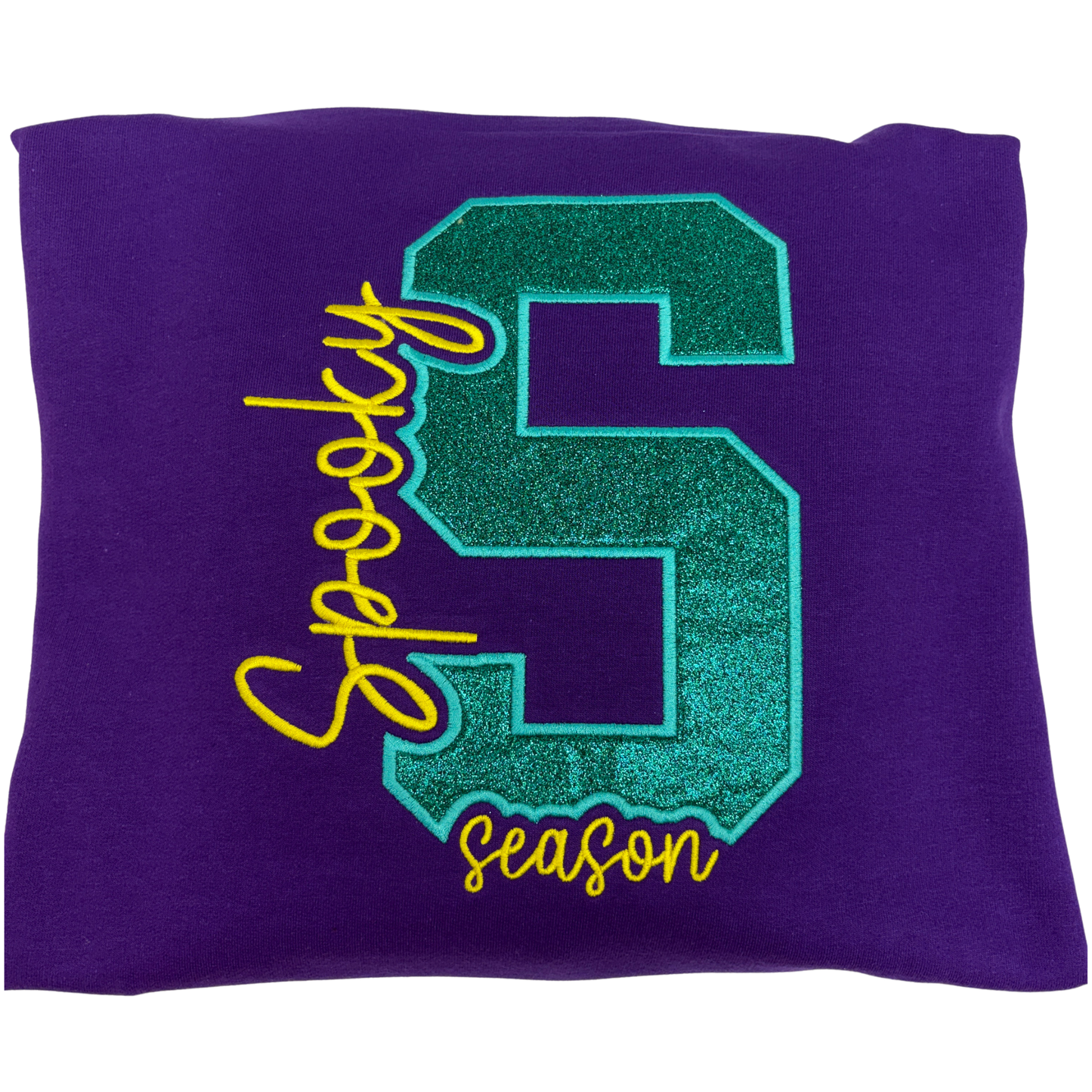 A purple sweatshirt featuring a large glittery teal collegiate letter S with the word 'Spooky on the left and season under it embroidered  in yellow thread