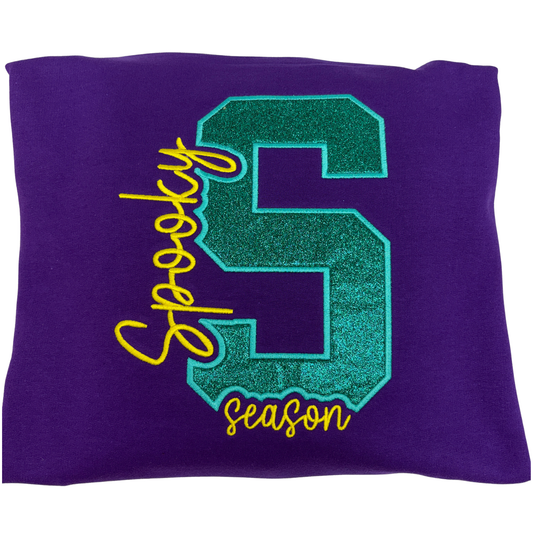A purple sweatshirt featuring a large glittery teal collegiate letter S with the word 'Spooky on the left and season under it embroidered  in yellow thread