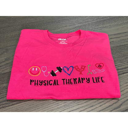 Folded pink t-shirt with 'Physical Therapy Life' text and symbols on a wooden surface