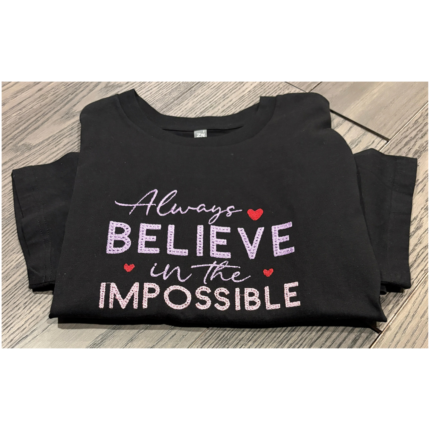 Black t-shirt with 'Always Believe in the Impossible' text on a wooden surface