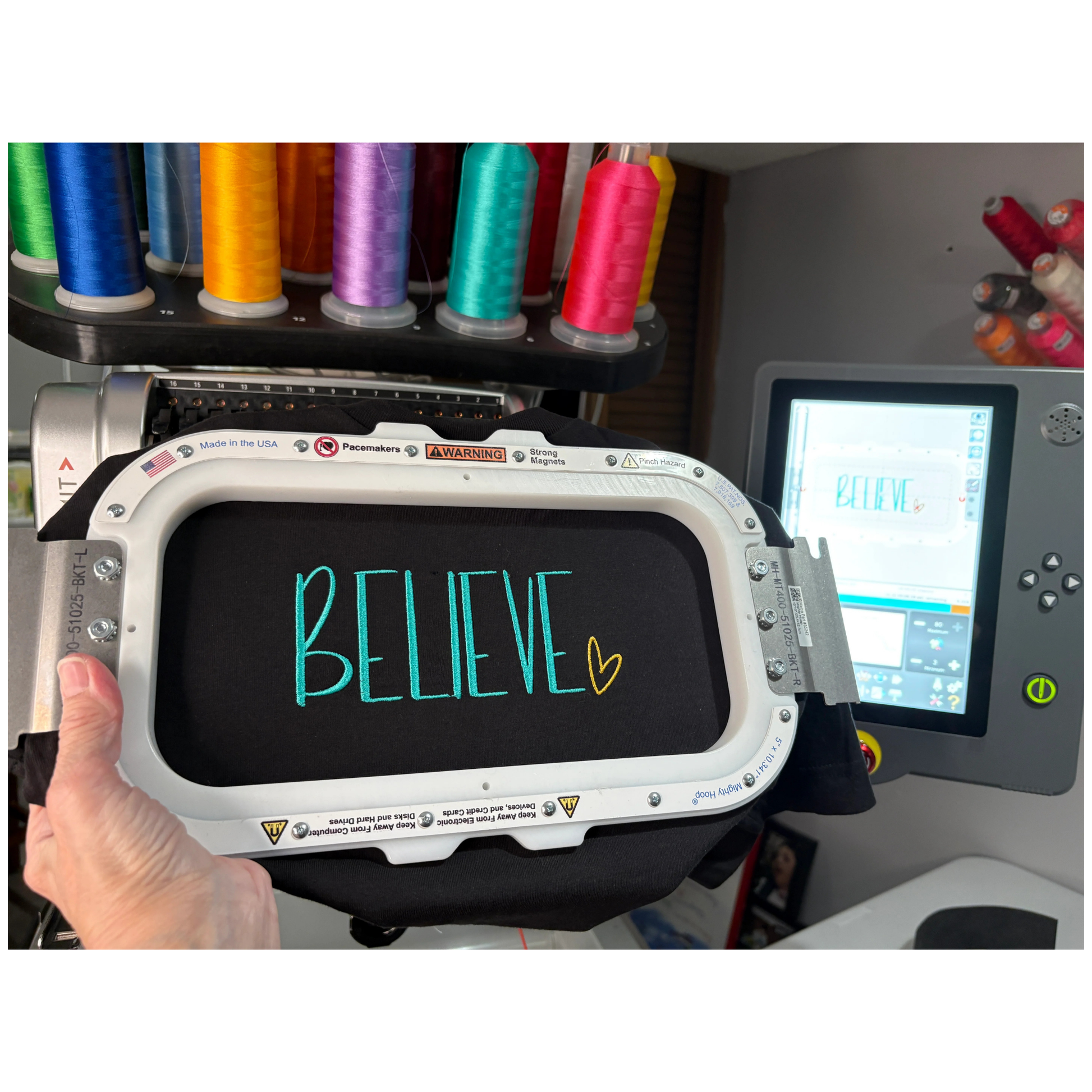 Embroidery machine with 'BELIEVE' text on a hoop, surrounded by colorful thread spools.