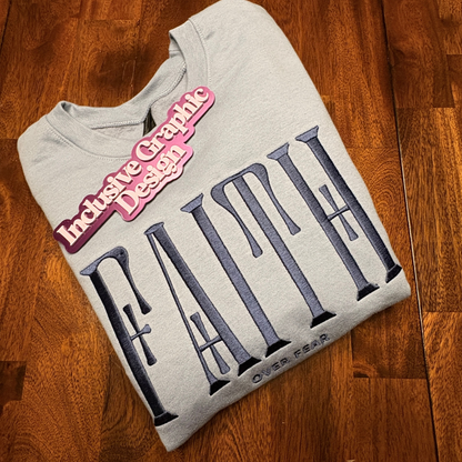A folded light gray sweatshirt with a large dark navy 'FAITH' graphic and the words 'over fear' beneath it. a pink 'Inclusive Graphic Design' magnet is displayed on top