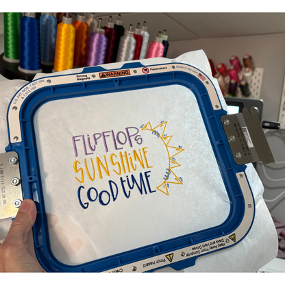 An embroidery hoop with a white fabric backing with an embroidered text reading 'FLIPFLOP SUNSHINE GOOD TIME' and a yellow sunburst design.  Spools of thread are blurred in the background.