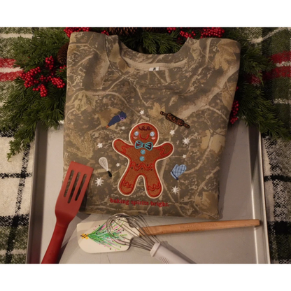 A camouflage-print sweatshirt laid flat on a metal tray, decorated with a gingerbread man graphic and the words 'Baking Spirits Bright.' Holiday greenery and red berries frame the top, and a red spatula, spatula scraper, and wooden whisk are placed in front.