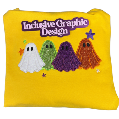 A folded yellow sweatshirt featuring an applique design of heat transfer vinyl of four glittery ghosts in white, green, purple, and orange, with a brand marker reading 'Inclusive Graphic Design' in purple.