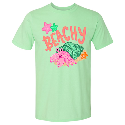 A mint green short-sleeve t-shirt with a colorful graphic on the front featuring the word 'BEACHY' in coral letters, pink and green starfish, and a pink hermit crab inside a green shell.