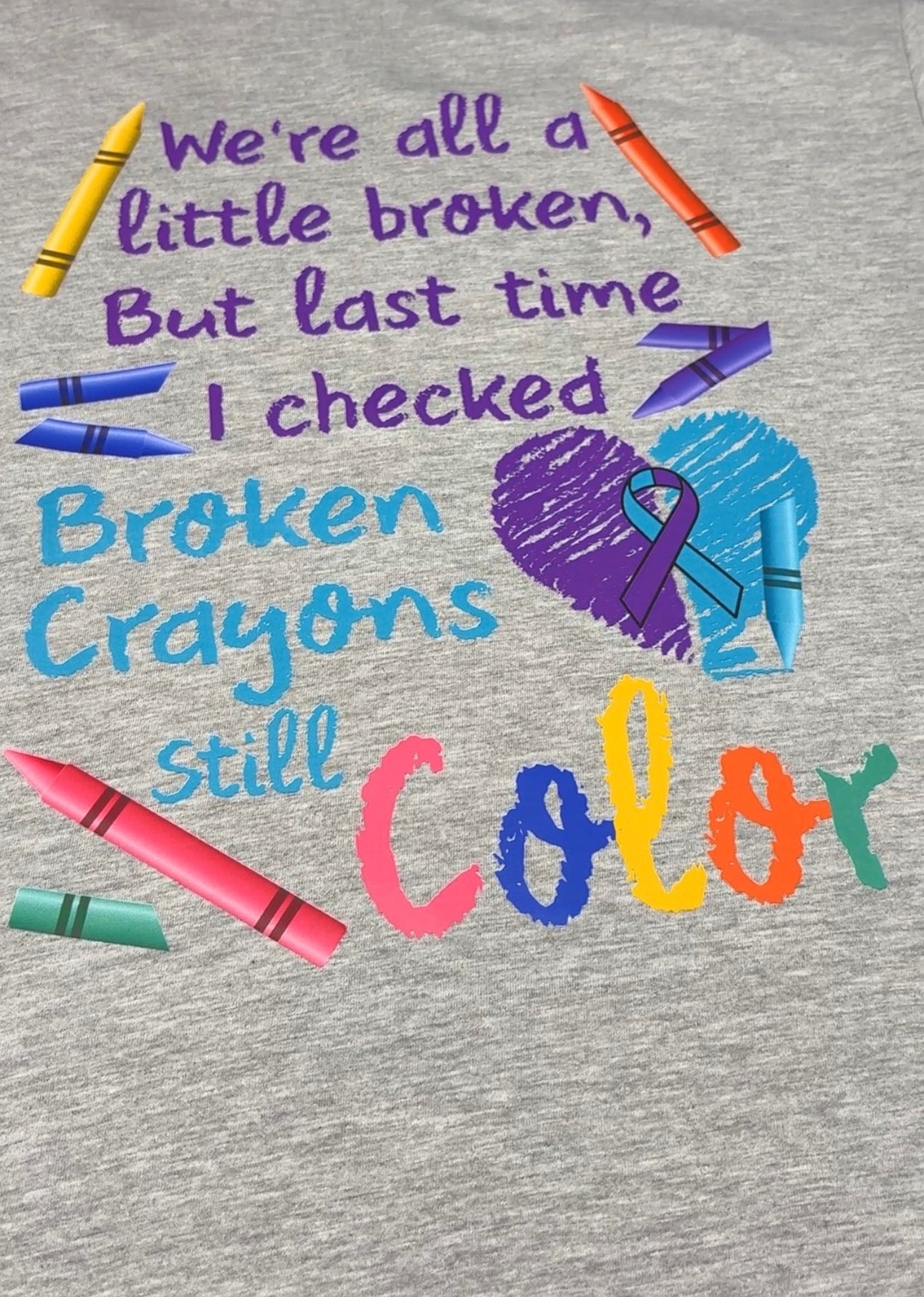 A close-up of a heather gray t-shirt featuring a colorful graphic design with the text 'We're all a little broken, But last time I checked Broken Crayons still Color' accompanied by illustrations of crayons and a purple awareness ribbon.