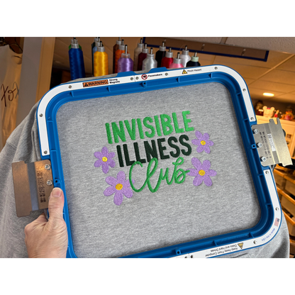 Embroidery hoop with 'Invisible Illness Club' text and flowers on a gray fabric.