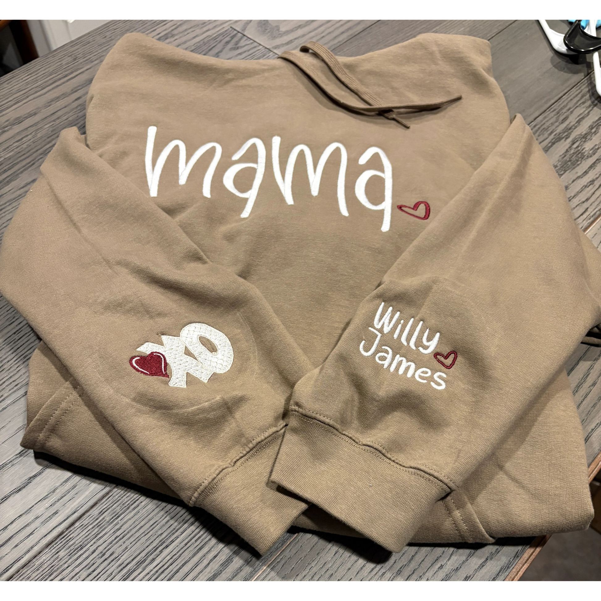 Folded brown hoodie with 'mama', 'Willy', and 'James' text on a wooden surface