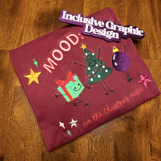 A  burgundy shirt featuring an embroidered Christmas-themed graphics, saying 'MOOD', cue the Christmas Music and a green Christmas tree, a purple ornament, a green gift box, and stars, placed on a wooden table with a purple 'Inclusive Graphic Design' magnet above it.