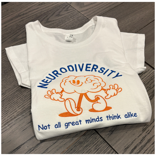 Folded white t-shirt with 'Neurodiversity' embroidered graphic and text that says No 2 minds think alike on a wooden surface