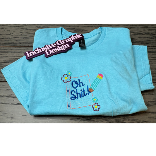 Light blue t-shirt with colorful graphic design customized notepad saying oh shit on a wooden surface, labeled 'Inclusive GraphicDesign'.