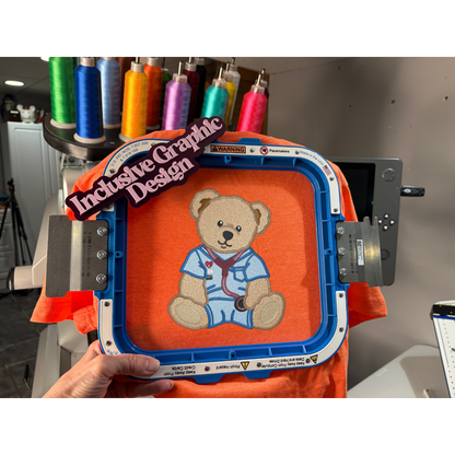 Embroidery hoop with teddy bear design on an orange fabric, held by a person in a workshop setting.