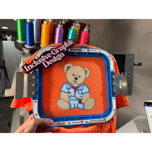Embroidery hoop with teddy bear design on an orange fabric, held by a person in a workshop setting.