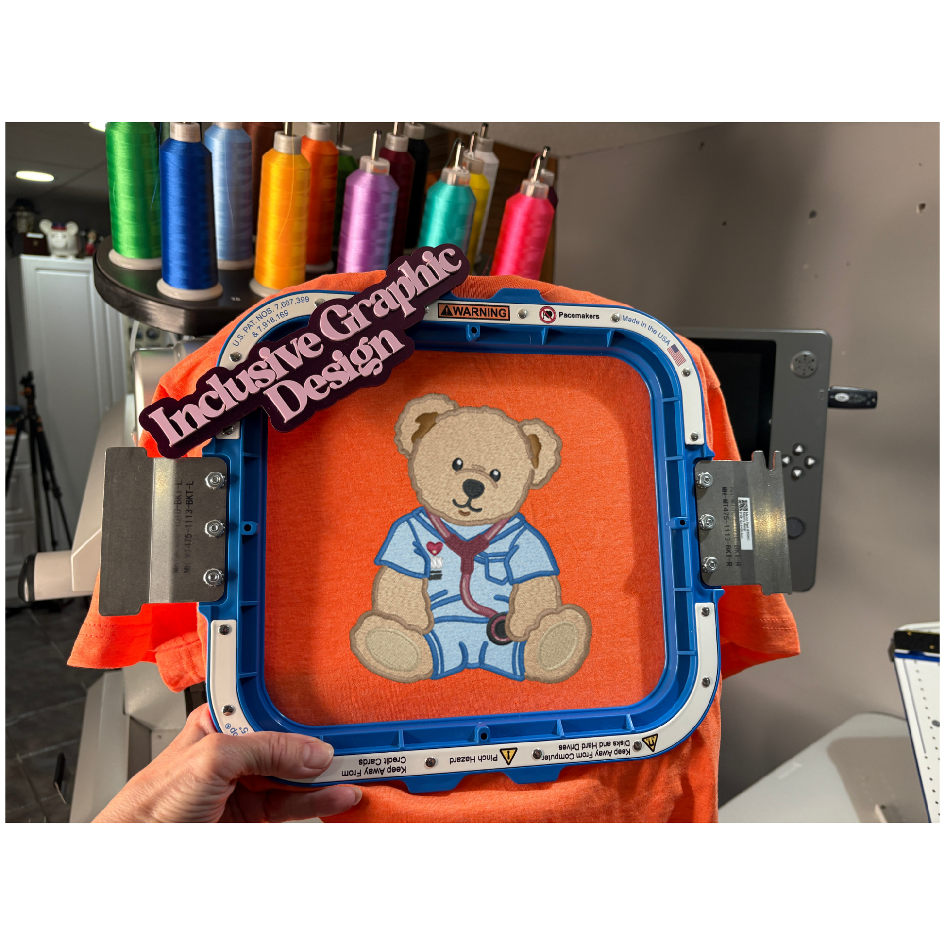 Embroidery hoop with a teddy bear design on an orange fabric, surrounded by spools of thread.