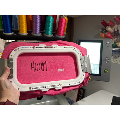 Pink embroidery machine hoop with 'Heart warrior mama' branding in an embroidery hoop with spools of thread in the background.