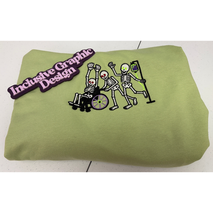A folded light olive green sweatshirt featuring a graphic of three dancing skeletons, one in a wheelchair, one pushing the skeleton in the wheelchair and another skeleton pulling an iv pole filled with green and purple fluid. A purple 'Inclusive Graphic Design' brand marker is on the sweatshirt.