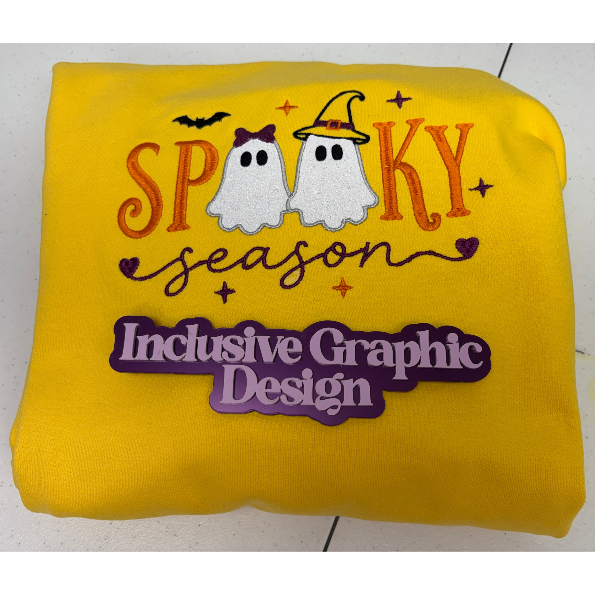 A bright yellow sweatshirt, featuring an appliquéd embroidered 'Spooky Season' design with two ghosts and the text 'Inclusive Graphic Design'.
