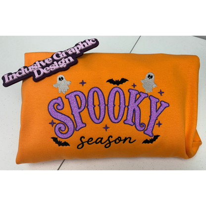 An orange sweatshirt and purple and black applique embroidered spooky season  with small ghost, bat, and star details. A purple tag reading 'Inclusive Graphic Design' is attached near the top left.