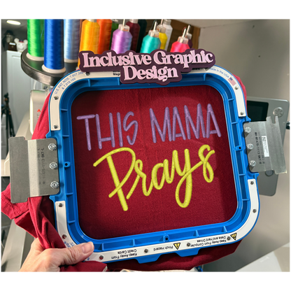 Embroidery hoop with 'This Mama Prays' text on a red fabric, surrounded by spools of thread.