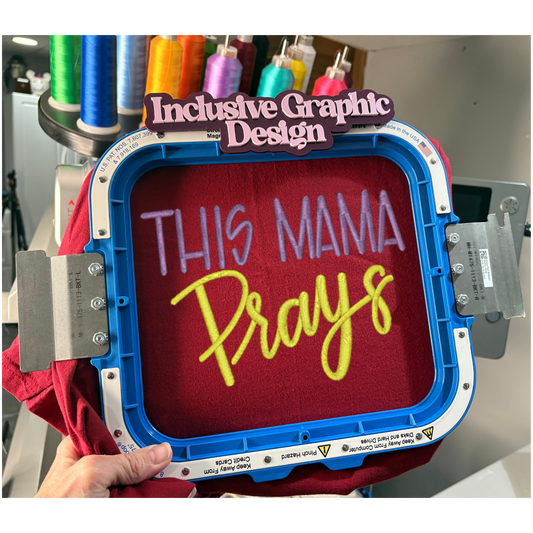 Embroidery hoop with 'This Mama Prays' text on a red fabric, surrounded by spools of thread.