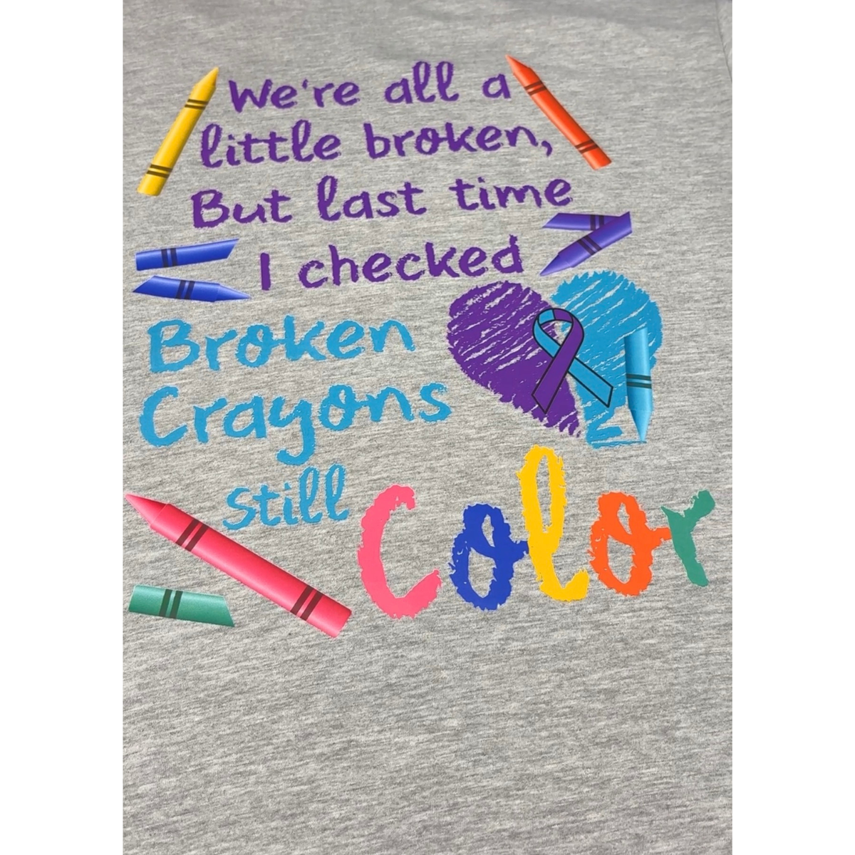 A close-up of a heather gray tshirt printed with the phrase 'We're all a little broken, But last time I checked Broken Crayons still Color' in purple and blue lettering, accompanied by illustrated crayons and a purple awareness ribbon.