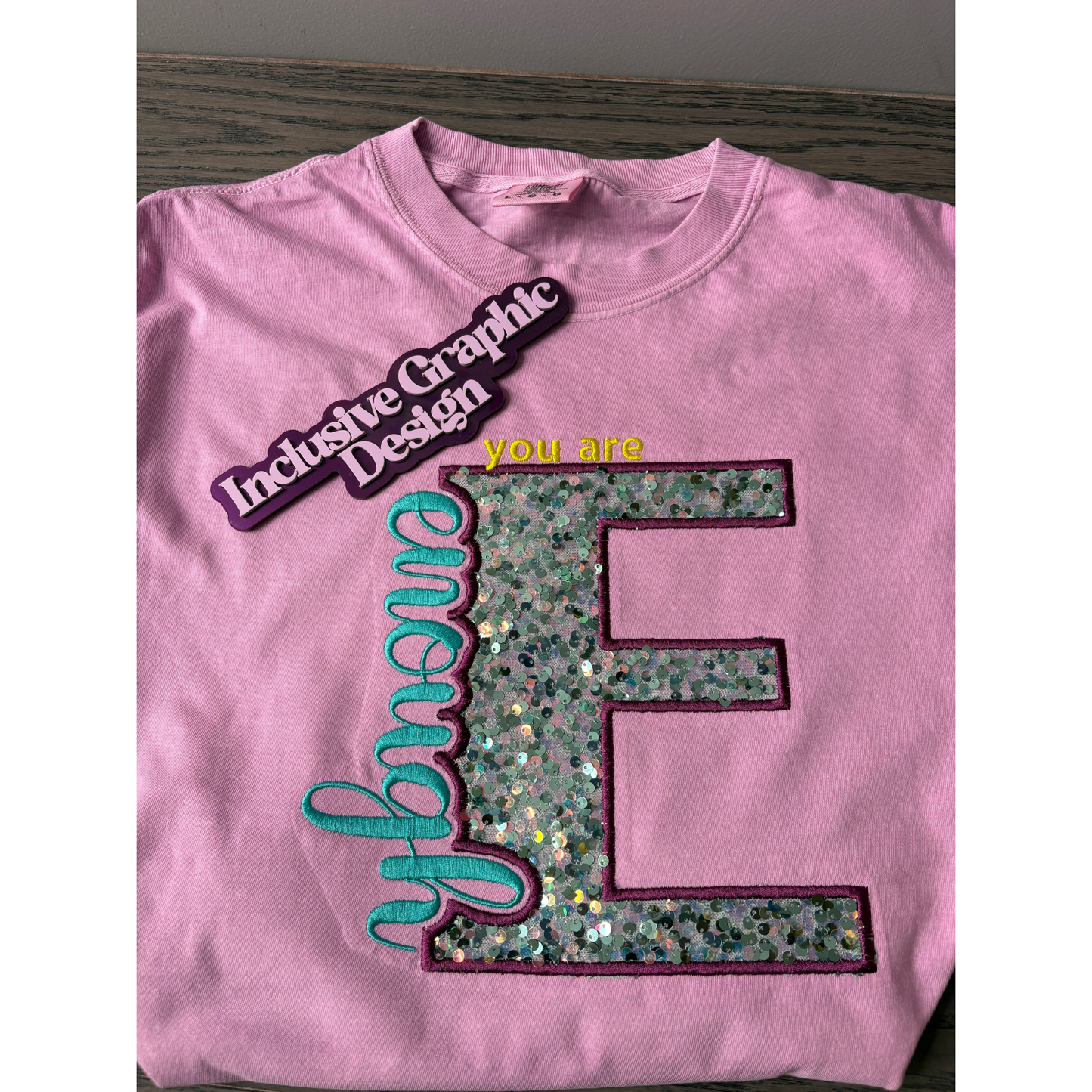 Pink Shirt with the letter E embroidered with sequence applique and the words You are enough and the water marker inclusive graphic design